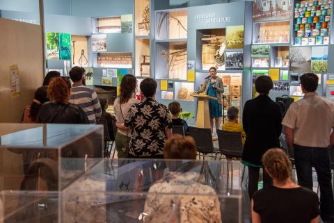 A group of people gathered in an exhibition space, some seated and others standing, facing a speaker at a podium. Behind the speaker is a wall display with images, artifacts, and the prominent text 'ABUNDANCE AGRICULTURE'. Glass display cases with items are visible in the foreground.