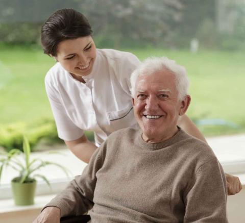 a caregiver stands over an elderly man in a wheel chair who is sitting in front of a window.