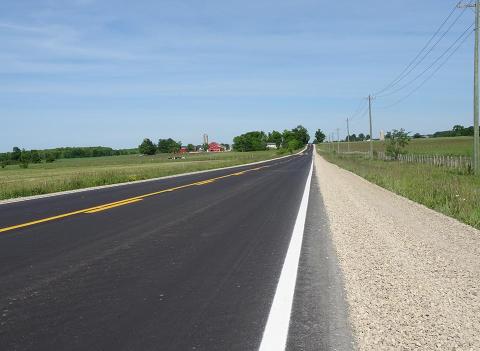 A freshly resurfaced Grey County road runs runs into the horizon. There is a farm with a large red barn in the distance.  Both sides of the roads are lined with hay fields.