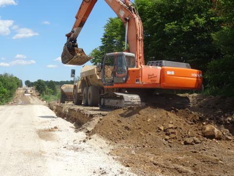 A high hoe scoops soil into the back of a dump truck on a County road