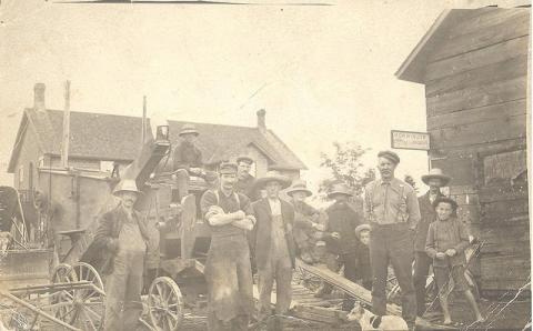 Black and white photograph with a group of people standing in the centre of a small village.  The picture looks to be quite old.