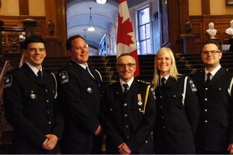 Five Grey County paramedics wearing their formal uniforms stand in front of a Canadian flag inside queen's park in Toronto.