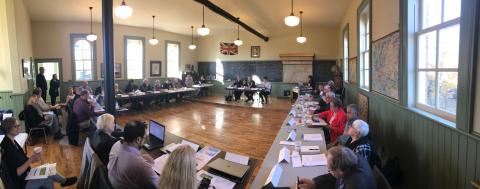 members of grey county council and county staff sit around a large horse shoe in the school house at Grey roots. The front wall is covered by a large chalk board and there are maps around the room.