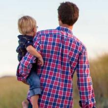 Photo of a father in a plaid shirt carrying a young, blonde haired toddler through a field.