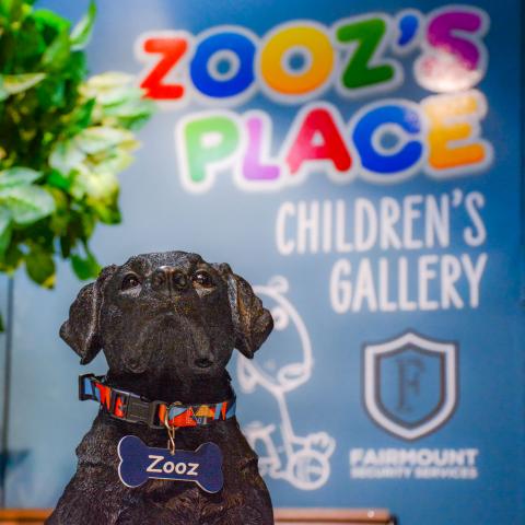 A dog wearing a nametag in front of text reading "Zooz's Place Children's Gallery"