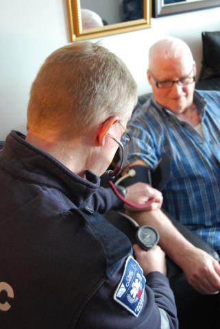 A community paramedic leans over an elderly man and checks his blood pressure.